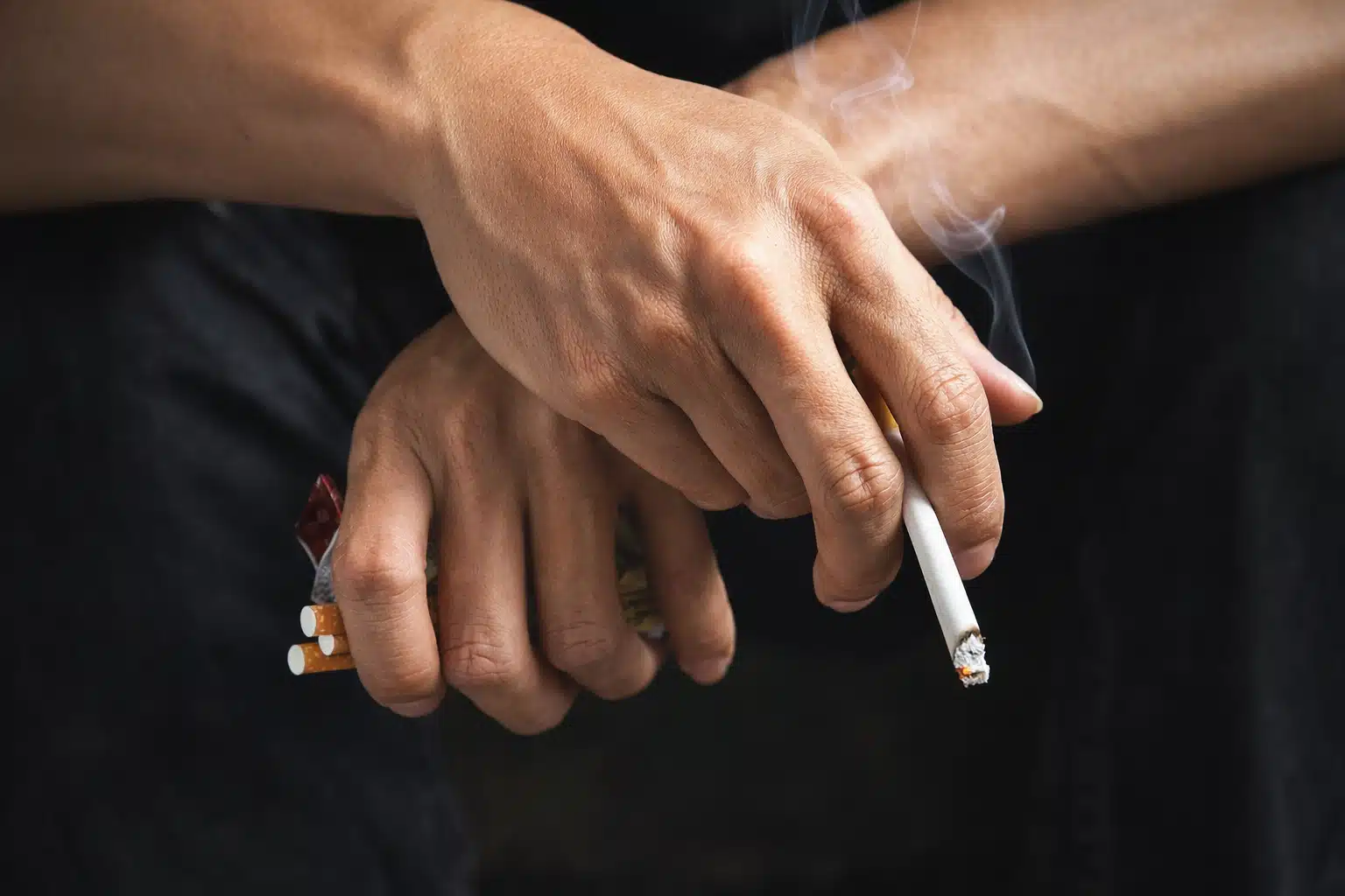 migliori rimedi per smettere di fumare dipendenza da nicotina Mani che tengono una sigaretta accesa e un pacchetto di sigarette, primo piano realistico sulla dipendenza da nicotina e sul gesto del fumo.